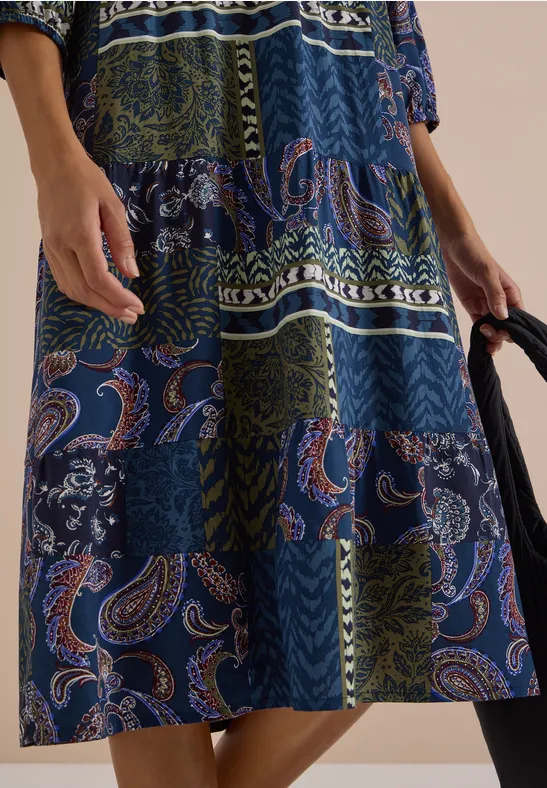 Robe patchwork mi-longue aux motifs cachemire et fleurs bleues et vertes, tenue avec un sac noir.