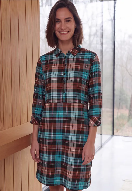 Une femme souriante porte une robe-chemise à carreaux devant un intérieur lumineux.