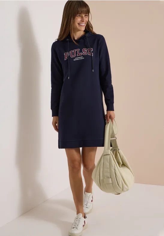 Femme portant une robe sweat à capuche bleue avec inscription, baskets blanches et grand sac.