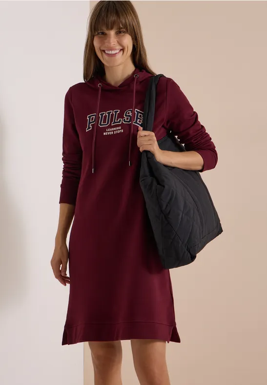 Femme en robe à capuche bordeaux avec l'inscription «Pulse», souriante, portant un grand sac noir.