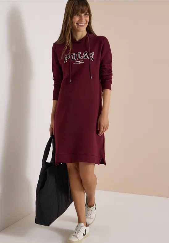 Femme en robe à capuche bordeaux portant l'inscription "Poler", sac noir à la main et baskets blanches.