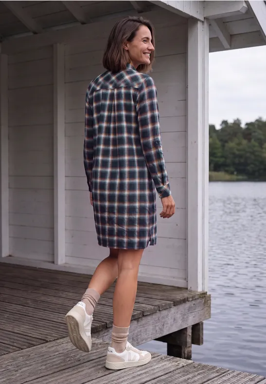 Femme en robe chemise à carreaux debout sur une jetée, regardant un lac paisible.