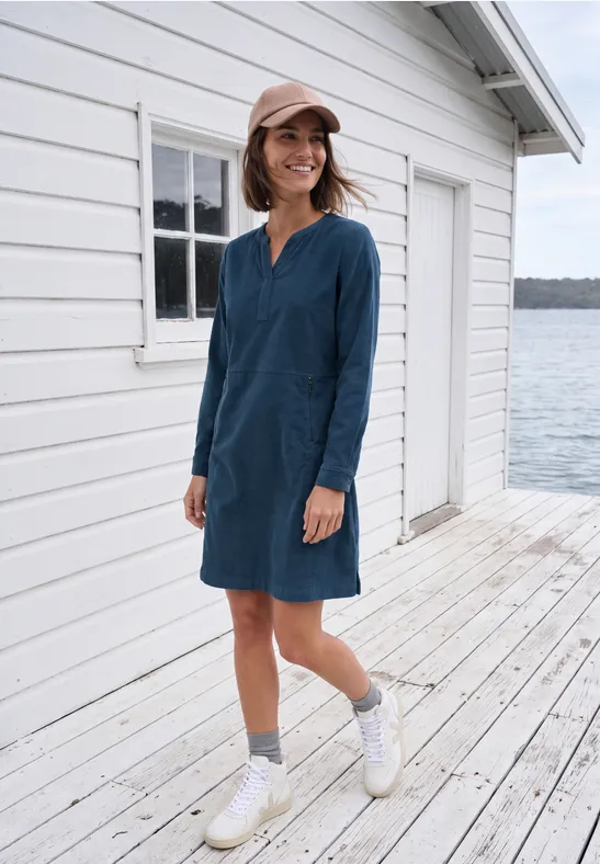 Femme en robe bleue à manches longues souriant sur une jetée en bois devant une maison blanche au bord de l'eau.