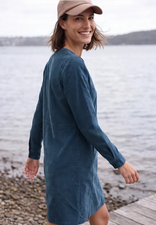 Une femme souriante en robe jean bleu et casquette, se tenant au bord de l'eau, regarde par-dessus son épaule.