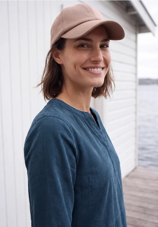 Femme souriante en casquette et haut bleu, debout au bord de l’eau devant un bâtiment blanc.