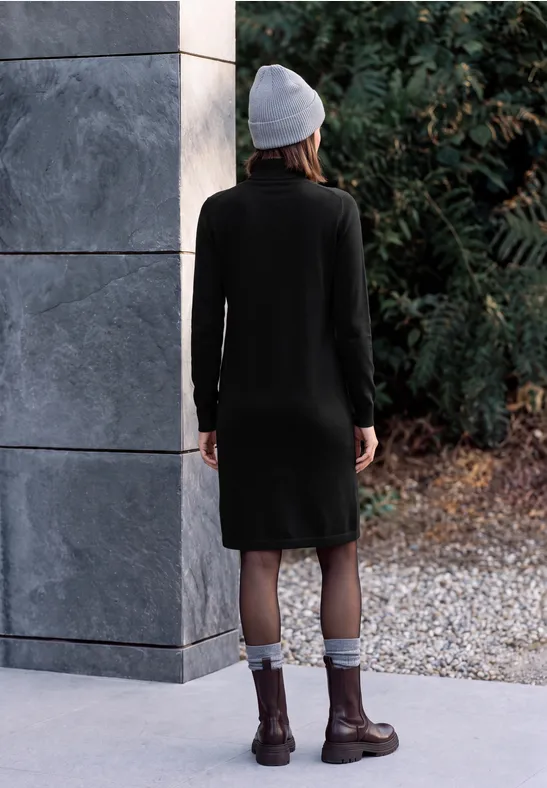 Une femme en robe noire, bottes et bonnet, vue de dos devant un décor naturel