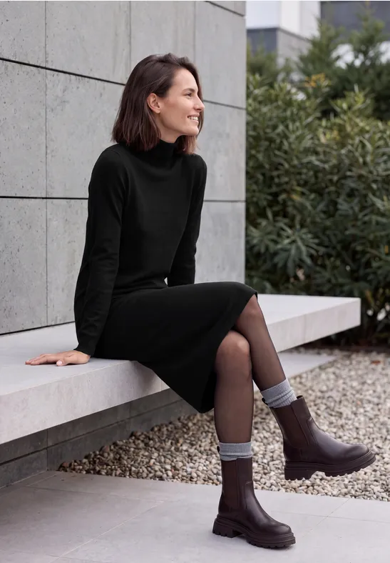 Femme en robe noire à col roulé, assise sur un banc extérieur, souriante, chaussettes grises et bottines marron.