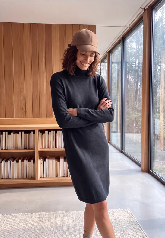 Frau in dunkelgrauem Kleid und Basecap lehnt lächelnd vor Regalen und großen Fenstern mit Blick ins Freie.