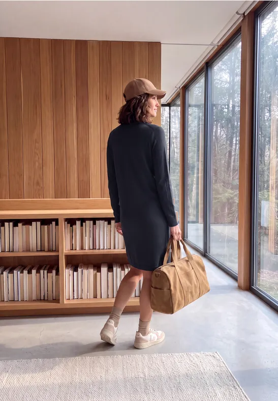 Eine Frau mit Mekko und Kappe steht in einem hellen Raum, hält eine Reisetasche, zwischen Bücherregalen und Fenstern.
