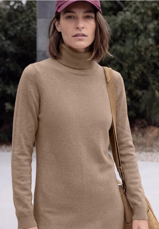 Une femme en pull à col roulé beige et casquette se tient dehors devant des buissons verts.