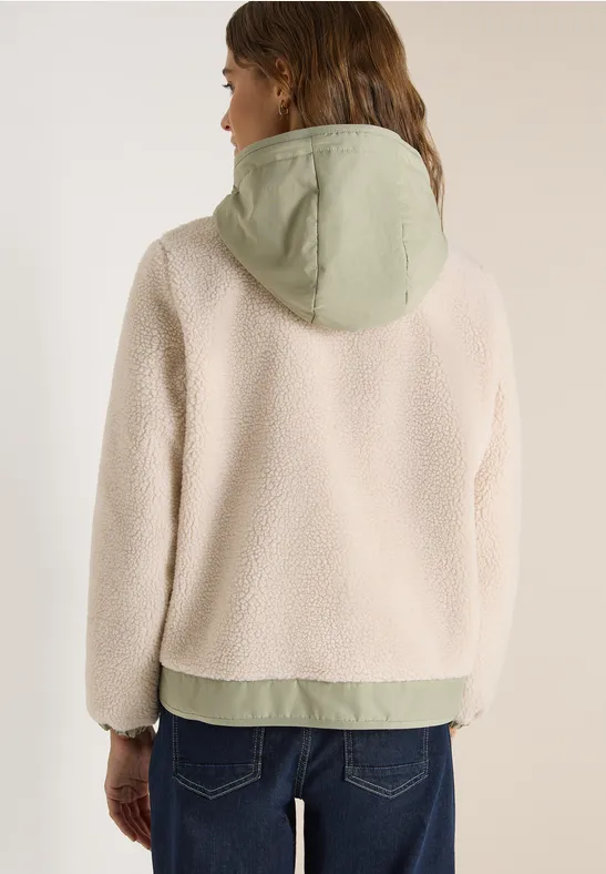 Rückenansicht: Person trägt cremefarbene Fleecejacke mit grünem Kapuzeneinsatz und blaue Jeans.