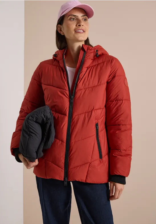 Frau in roter Steppjacke mit Kapuze, trägt rosa Cap und hält schwarze, zusammengefaltete Jacke, lächelt.