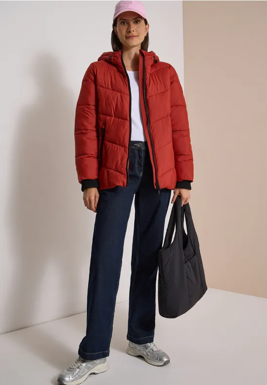 Frau in roter Steppjacke, weißem Shirt, blauen Jeans und Sneakern trägt pinke Kappe und schwarze Tasche.