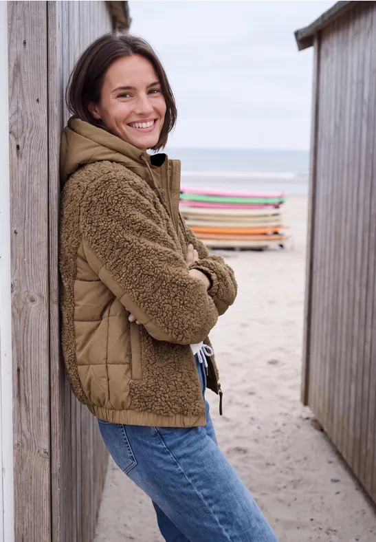 Eine junge Frau lehnt lächelnd zwischen Strandhütten am Meer.