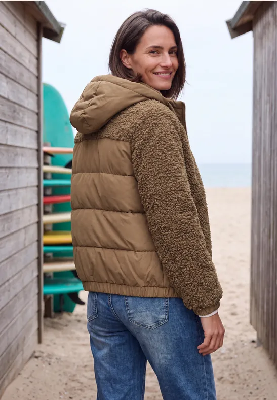 Frau in brauner Stepp- und Fleecejacke steht zwischen Strandhütten und blickt lächelnd zurück.