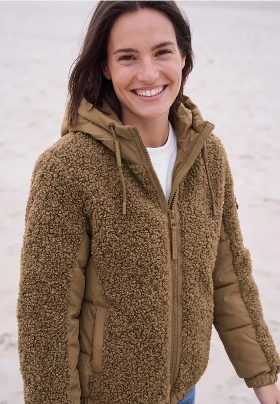 Frau lächelt am Strand in brauner Teddy-Fleece-Jacke mit Kapuze.
