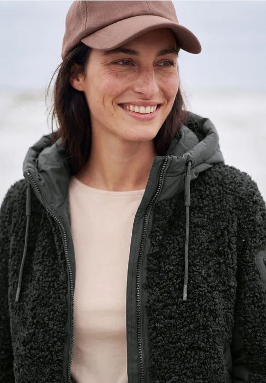 Eine lächelnde Frau mit Basecap und kuscheliger Fleece-Jacke am Strand.