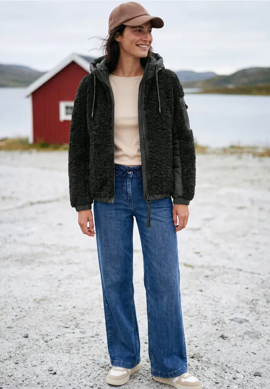 Frau in schwarzer Teddyjacke, weiten Jeans und Cap steht vor rotem Haus am See in ruhiger Landschaft.