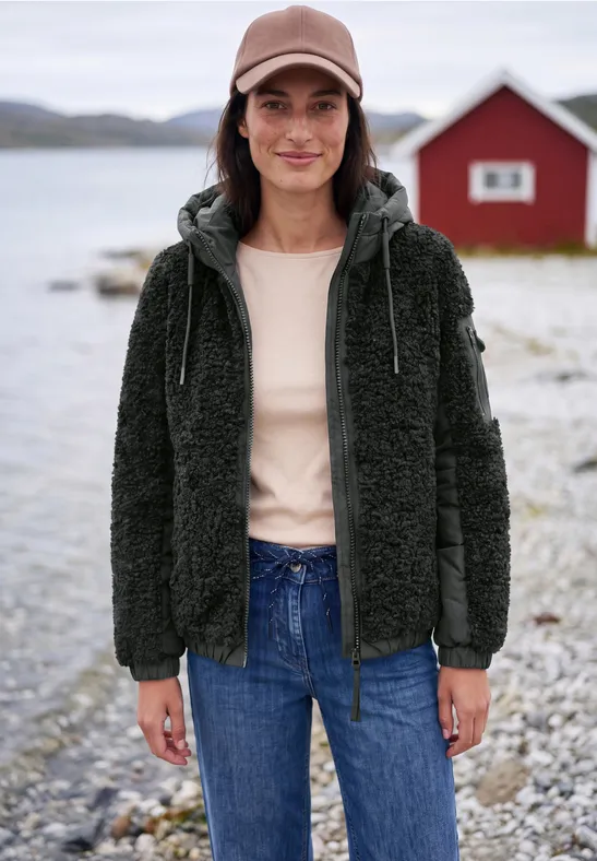 Frau in schwarzer Teddyjacke und Jeans steht am kiesigen Ufer mit rotem Haus im Hintergrund.