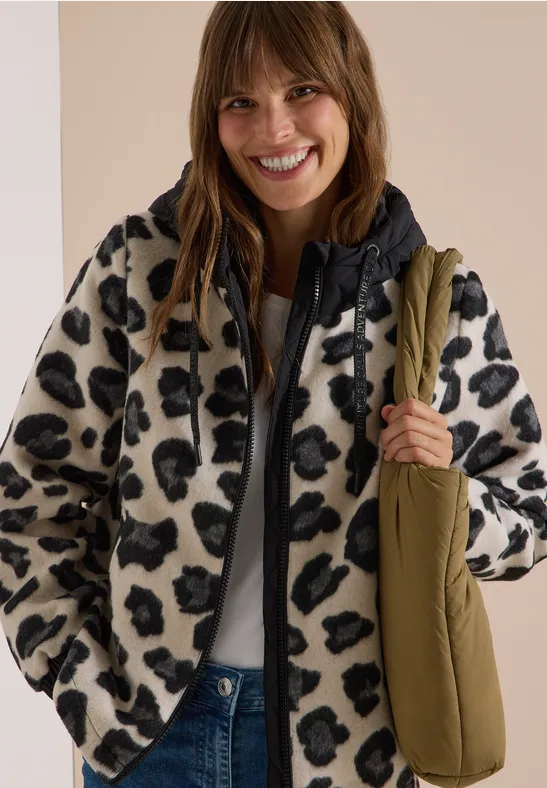 Lächelnde Frau in Teddyjacke mit Leopardenmuster und olivgrüner Umhängetasche.