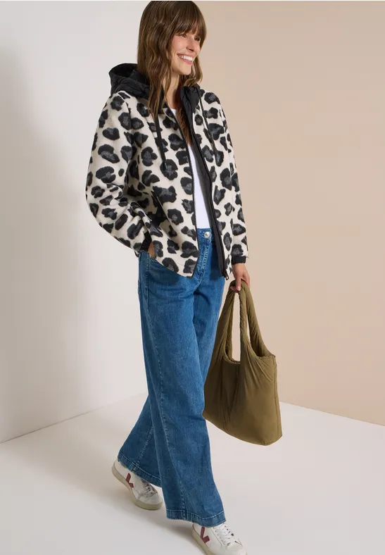 Frau in modischer, bedruckter Jacke, weitem Jeans, weißen Sneakers und grünem Shopper vor neutralem Hintergrund.