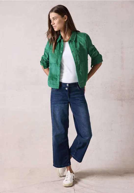 Frau mit grüner Jacke, weißem Shirt, blauer Jeans und Sneakern steht lässig.