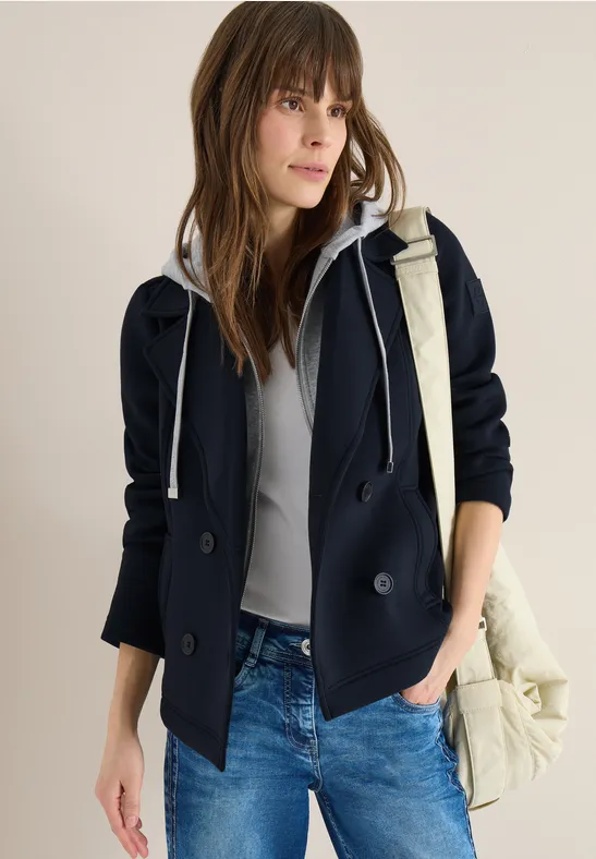 Lässiger Look: dunkelblauer Kapuzenblazer, hellgraues Shirt, blaue Jeans und beige Tasche vor neutralem Hintergrund.