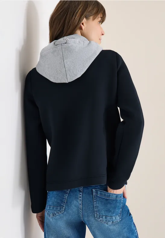Rückansicht einer Person in schwarzem Pullover mit grauer Kapuze, Hände in den Jeans-Taschen, lehnt an Wand.