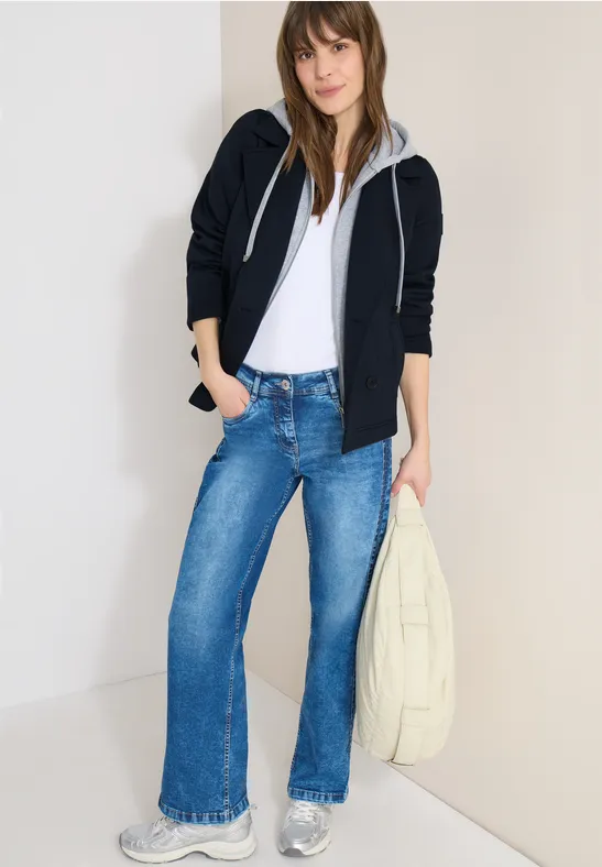 Frau trägt weißes T-Shirt, schwarze Kapuzenjacke, weite blaue Jeans und helle Stofftasche.