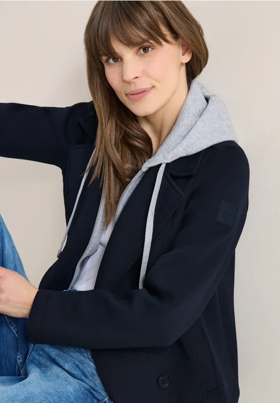 Frau trägt grauen Kapuzenpullover unter dunkelblauem Blazer und Jeans.