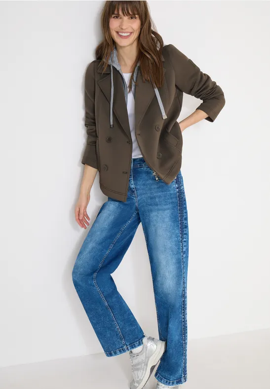 Frau trägt olivgrüne Jacke, grauen Hoodie, blaue Jeans und weiße Sneaker in legerer Pose.