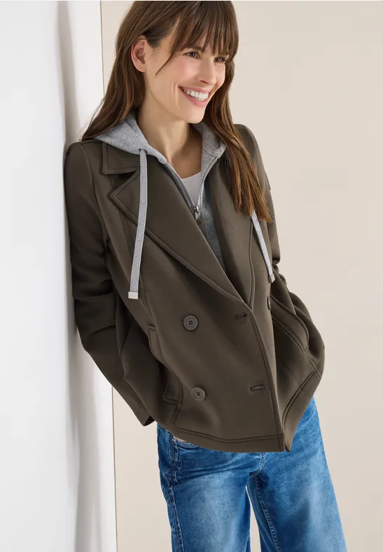 Dame lehnt in schwarzem Blazer, grauem Kapuzenpullover und blauen Jeans an weißer Wand.