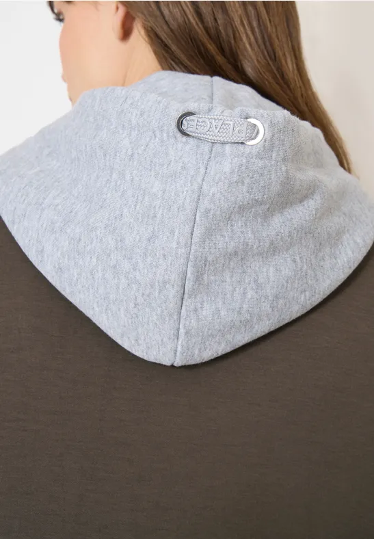 Nahaufnahme einer Person von hinten in braun-grauer Kapuzenjacke mit silbernem Logo-Patch am Kapuzenansatz.