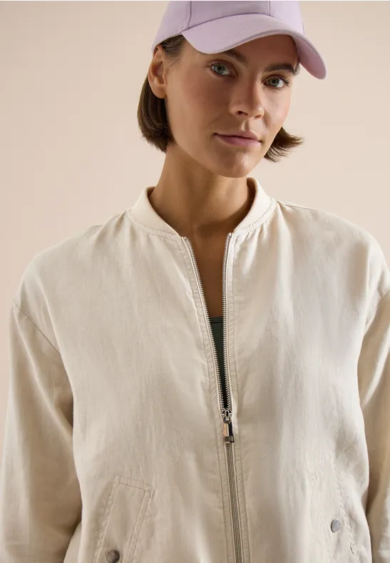 Frau in hellbeiger Jacke mit Reißverschluss und rosa Baseballcap vor neutralem Hintergrund.