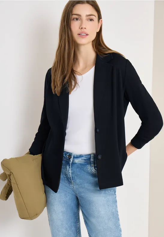 Frau in schwarzer Strickjacke, weißem Shirt, blauen Jeans, hält eine olivgrüne Tasche.