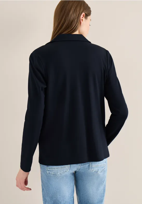 Eine Frau steht mit dem Rücken zur Kamera, trägt einen dunkelblauen Blazer und hellblaue Jeans vor beigem Hintergrund.