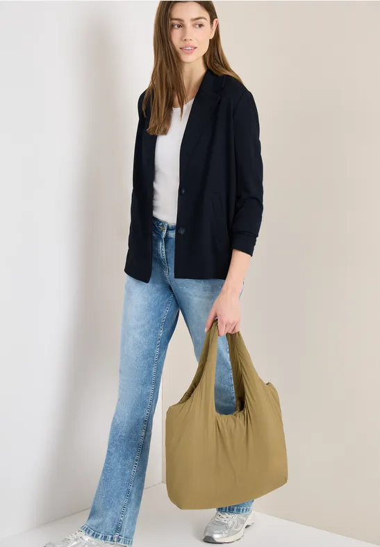 Frau trägt weiße Bluse, schwarze Strickjacke, Schlagjeans, weiße Sneaker und hält große grüne Tasche.