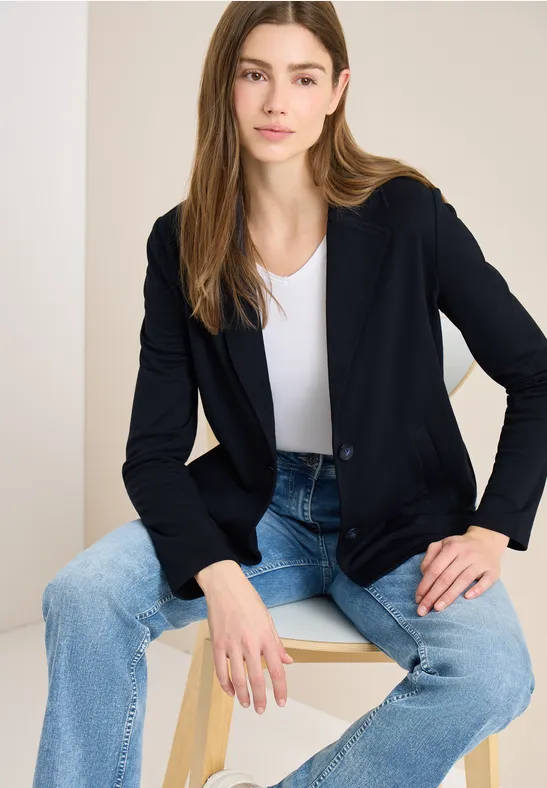 Eine Person sitzt auf einem Hocker, trägt weiße Bluse, schwarze Jacke und blaue Jeans.