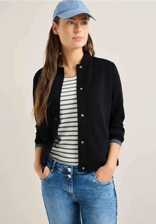 Frau in lässiger Kleidung: marineblaue Strickjacke, gestreiftes T-Shirt, blaue Jeans und hellblaue Baseballmütze.