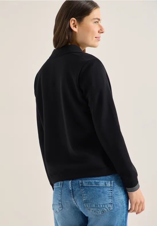 Person in schwarzem Pullover und blauen Jeans steht seitlich vor hellem Hintergrund.