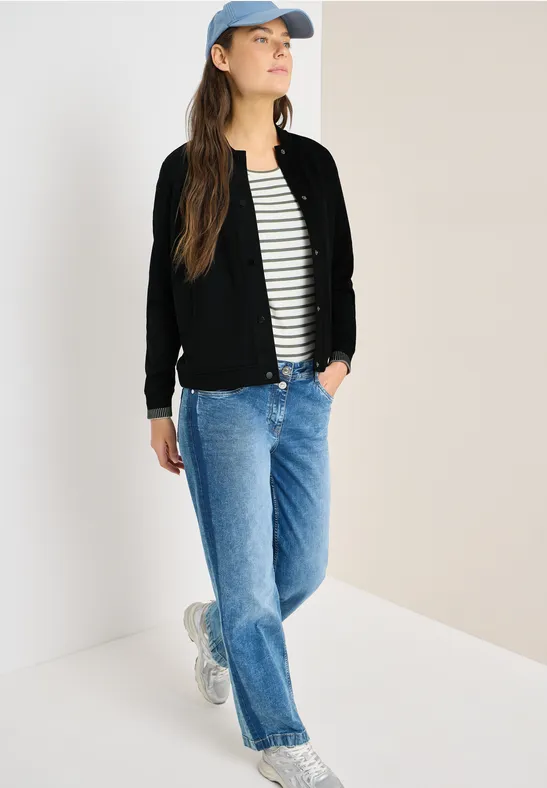 Person in schwarzer Jacke, gestreiftem Shirt, blauen Jeans, Sneakern und Baseballcap vor hellem Hintergrund.