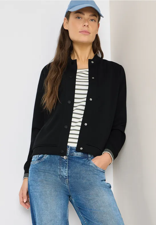 Frau in schwarzem Cardigan, gestreiftem Shirt und blauen Jeans vor hellem Hintergrund.