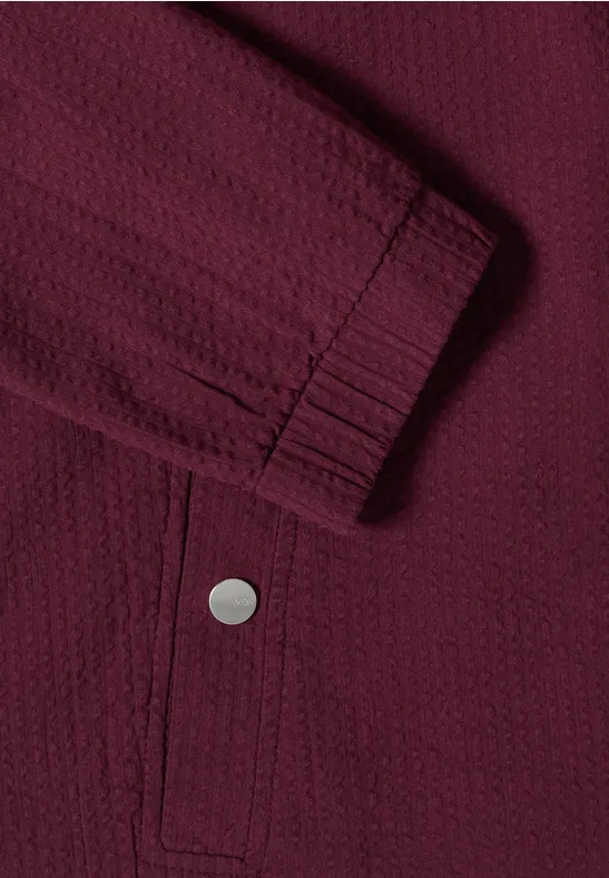 Nahaufnahme einer dunkelroten Jacke mit strukturiertem Stoff, elastischem Ärmelbündchen und Druckknopf-Tasche.