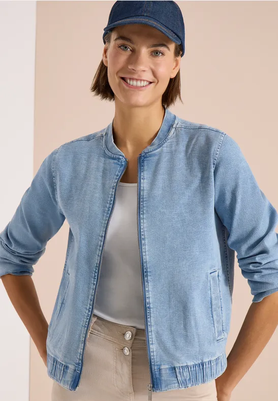 Eine Person trägt eine hellblaue Jeansjacke mit Reißverschluss, weißes Shirt und beige Hose vor hellem Hintergrund.