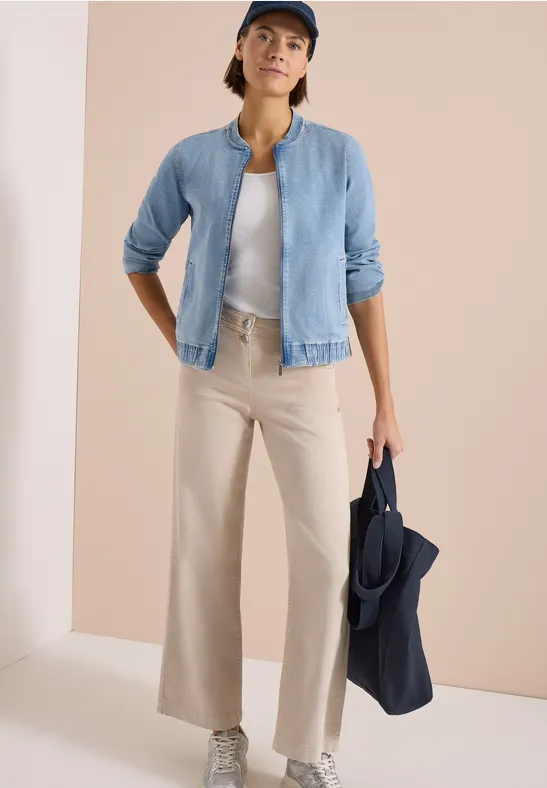 Dame im hellblauen Blouson, weißem Top und beigen Weite-Bein-Hosen, hält navy Blazer in heller Studio-Kulisse.