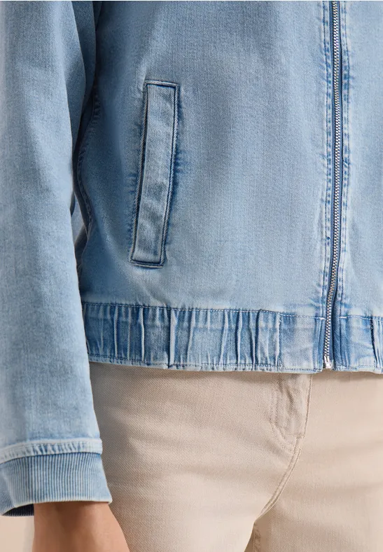 Leichtblaue Jeansjacke mit Reißverschluss und Seitentaschen über beigen Hosen.