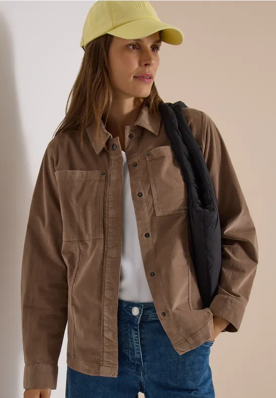 Eine Frau trägt eine beige Cordjacke, weißes Shirt, blaue Jeans, gelbe Kappe und schwarze Schultertasche.