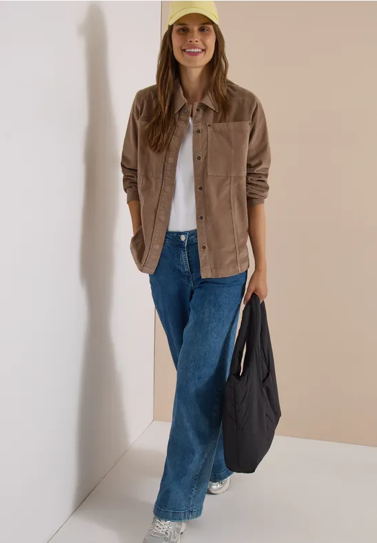 Eine lächelnde Frau mit gelber Mütze, beiger Cordjacke, Jeans und Tasche lehnt fröhlich an einer Wand.