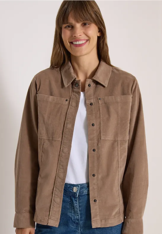 Eine Frau lächelt in einer braunen Cordjacke, weißem Shirt und blauen Jeans vor hellem Hintergrund.