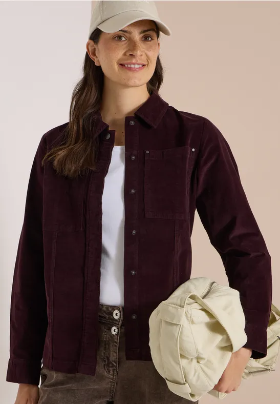 Frau in bordeauxfarbener Cordjacke, weißem T-Shirt und Cargohose hält lässig eine cremefarbene Tasche.